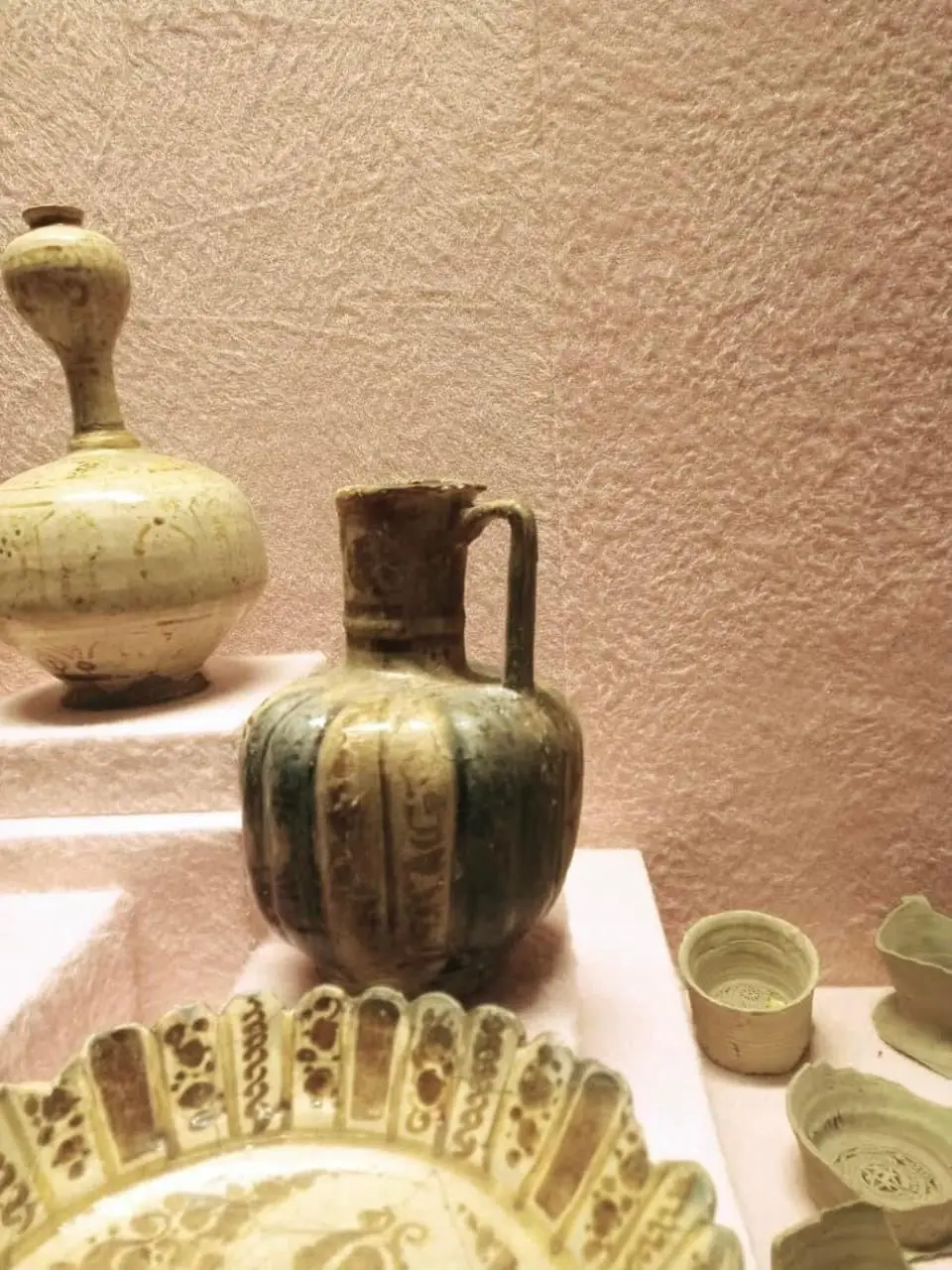 Glazed Islamic pottery in a museum display case: a ribbed green-and-ochre jug with a handle, a tall cream-glazed bottle, a decorated dish and small bowls. Published by Youm7 with its report on the Hurghada Antiquities Museum.