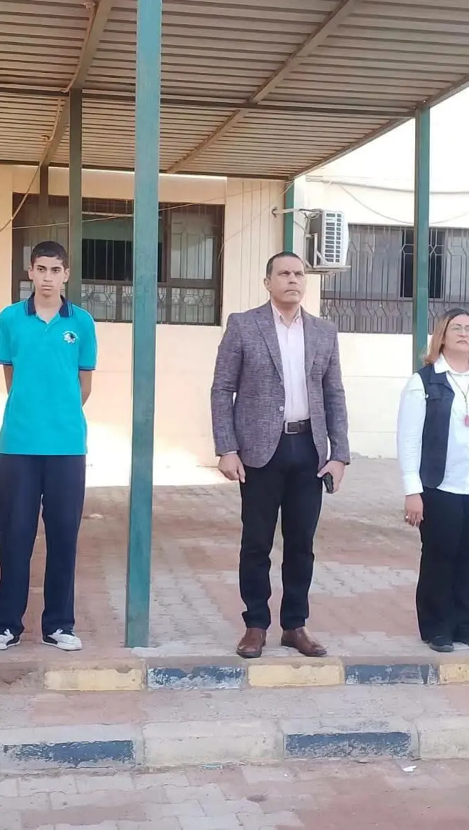 Five people, among them a pupil in school uniform, stand in a school yard under a shaded walkway during a morning assembly, one man speaking into a microphone. Photograph published by Al-Masry Al-Youm with its report on the inspection order; the individuals are not identified in the report.